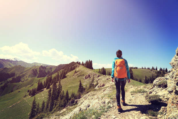 A person with an orange backpack hikes along a rugged mountain trail, surrounded by green hills and distant trees under a clear, blue sky.